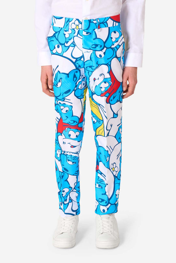 Een tienerjongen draagt ​​een opvallend Smurfenpak met kleurrijke cartoonfiguurprints in de kleuren blauw, rood, geel en wit, gecombineerd met een bijpassende stropdas en een wit overhemd, lachend tegen een effen achtergrond. Close up van de broek.