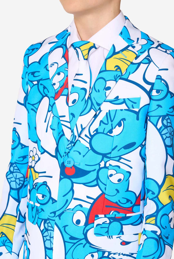 Een tienerjongen draagt ​​een opvallend Smurfenpak met kleurrijke cartoonfiguurprints in de kleuren blauw, rood, geel en wit, gecombineerd met een bijpassende stropdas en een wit overhemd, lachend tegen een effen achtergrond. Close up van de borst.
