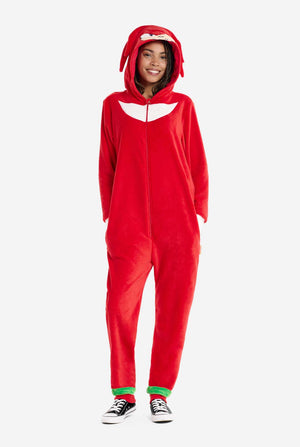 Een vrouw draagt ​​een felrode Knuckles the Echidna onesie voor volwassenen, een gezellige en speelse outfit geïnspireerd op het beroemde personage uit de Sonic the Hedgehog-serie. De onesie is voorzien van de kenmerkende witte borstpatch van Knuckles en heeft een capuchon die het gezicht van Knuckles laat zien, compleet met zijn expressieve ogen en lange rode dreads. De ritssluiting aan de voorkant zorgt voor gemakkelijk dragen en de vrouw staat met haar handen in de zijzakken.  