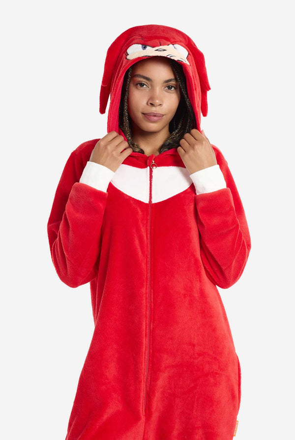 Een vrouw draagt ​​een felrode Knuckles the Echidna onesie voor volwassenen, een gezellige en speelse outfit geïnspireerd op het beroemde personage uit de Sonic the Hedgehog-serie. De onesie is voorzien van de kenmerkende witte borstpatch van Knuckles en heeft een capuchon die het gezicht van Knuckles laat zien, compleet met zijn expressieve ogen en lange rode dreads. De ritssluiting aan de voorkant zorgt voor gemakkelijk dragen, en heeft de capuchon op en houdt het vast.