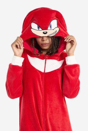 Een vrouw draagt ​​een felrode Knuckles the Echidna onesie voor volwassenen, een gezellige en speelse outfit geïnspireerd op het beroemde personage uit de Sonic the Hedgehog-serie. De onesie is voorzien van de kenmerkende witte borstpatch van Knuckles en heeft een capuchon die het gezicht van Knuckles laat zien, compleet met zijn expressieve ogen en lange rode dreads. De ritssluiting aan de voorkant zorgt voor gemakkelijk dragen, en heeft de capuchon op.