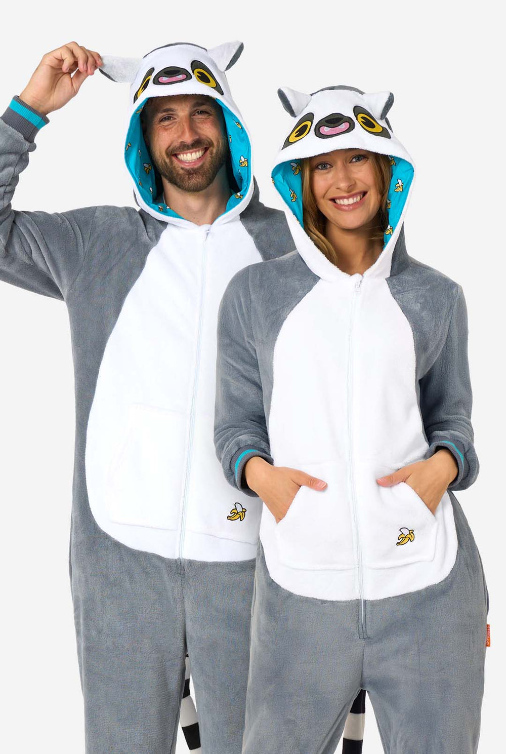Een man en vrouw dragen een grijze en witte maki onesie voor volwassenen met speelse capuchondetails en knus, zacht materiaal, perfect om in te loungen of voor verkleedpartijen. Inclusief zwart-witte staart. De capuchon staat op hun hoofd.