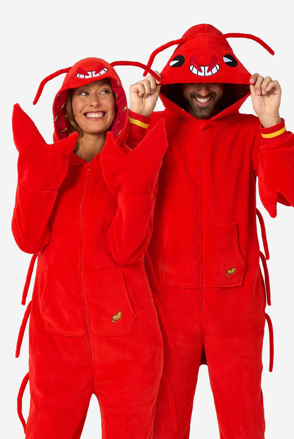 Een man en een vrouw dragen een felrode onesie voor volwassenen met kreeftenthema, speelse capuchondetails en knus, zacht materiaal, perfect om in te loungen of als verkleedkleding. Beiden dragen hun capuchon op hun hoofd.