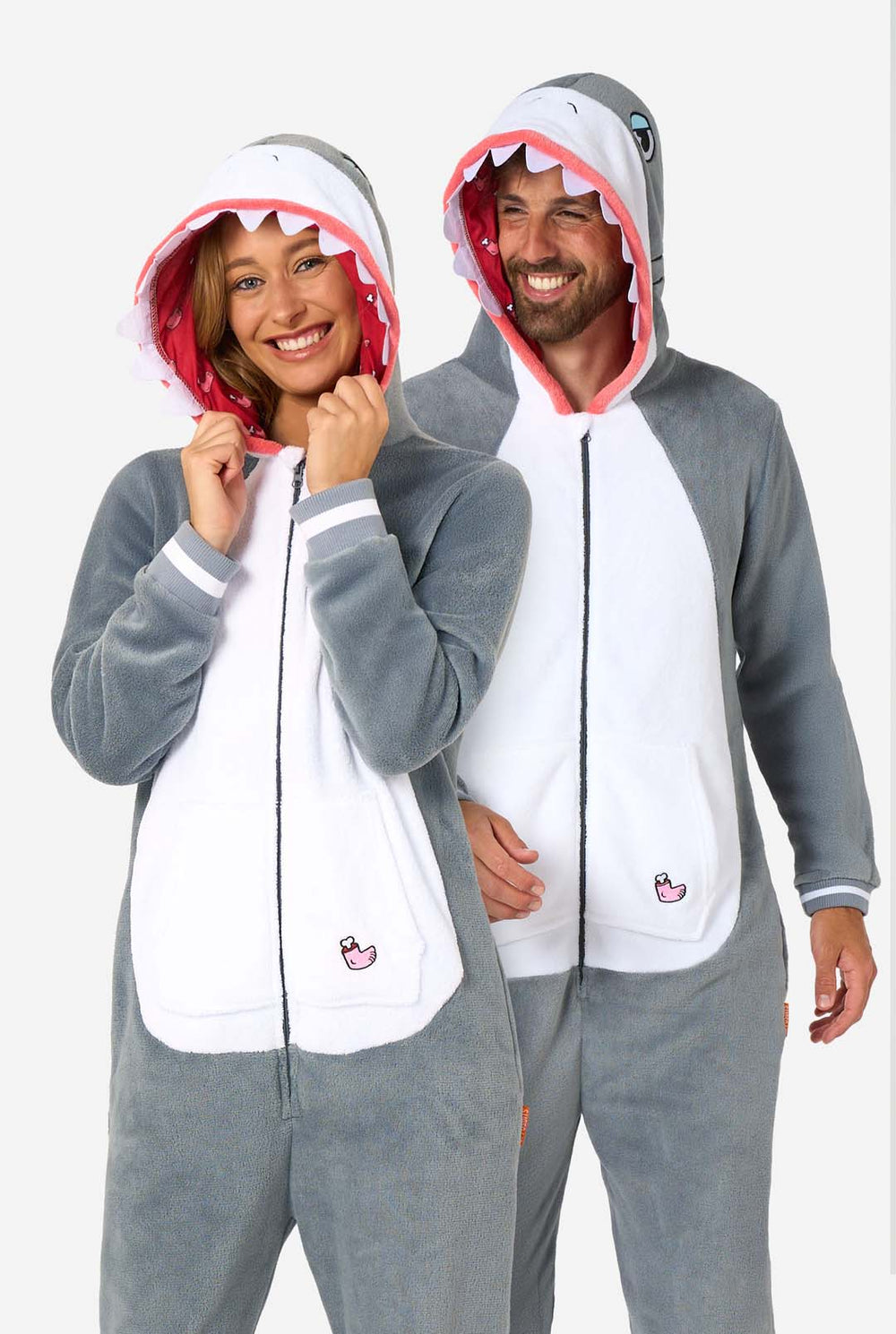 Een man en een vrouw dragen een grijs met witte haai onesie voor volwassenen met speelse details in de capuchon en knus, zacht materiaal, perfect voor loungen of verkleedpartijen. Beiden dragen hun capuchon op hun hoofd.