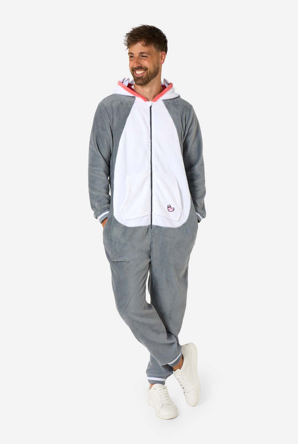 Een man draagt ​​een grijze en witte haaienonesie voor volwassenen met speelse capuchondetails en gezellig, zacht materiaal, perfect voor loungen of verkleedevenementen. Volledig lichaamsbeeld en twee handen in de zakken.