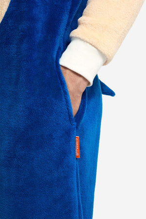 Een persoon is gekleed in een gezellige, blauwe Sonic the Hedgehog onesie voor volwassenen, met zachtbeige patches op de voorkant en mouwen om de iconische look van Sonic na te bootsen. De onesie heeft een capuchon ontworpen met het herkenbare gezicht van Sonic. De ritssluiting aan de voorkant zorgt voor een comfortabele pasvorm en de persoon staat met haar handen in de zijzakken van de onesie. 
