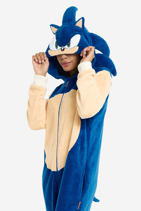 Een vrouw is gekleed in een gezellige, blauwe Sonic the Hedgehog onesie voor volwassenen, met zachtbeige patches op de voorkant en mouwen om de iconische look van Sonic na te bootsen. De onesie heeft een capuchon ontworpen met het herkenbare gezicht van Sonic. De ritssluiting aan de voorkant zorgt voor een comfortabele pasvorm. Haar uitdrukking is kalm en ontspannen. 