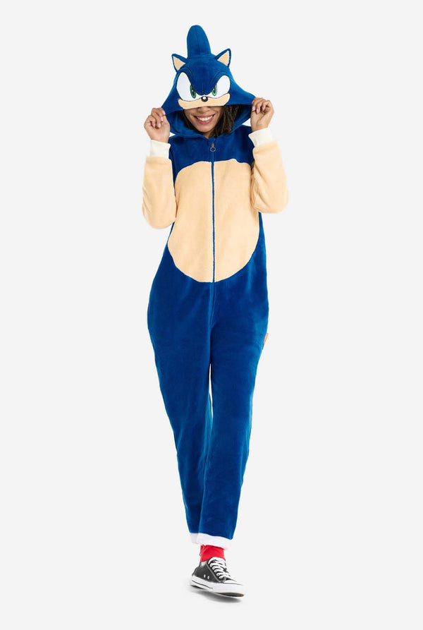 Een vrouw is gekleed in een gezellige, blauwe Sonic the Hedgehog onesie voor volwassenen, met zachtbeige patches op de voorkant en mouwen om de iconische look van Sonic na te bootsen. De onesie heeft een capuchon die is ontworpen met het herkenbare gezicht van Sonic, inclusief grote groene ogen, blauwe punten en puntige oren. De ritssluiting aan de voorkant zorgt voor een comfortabele pasvorm en de vrouw houdt de capuchon over haar ogen. Haar uitdrukking is kalm en ontspannen. Deze Sonic the Hedgehog onesie