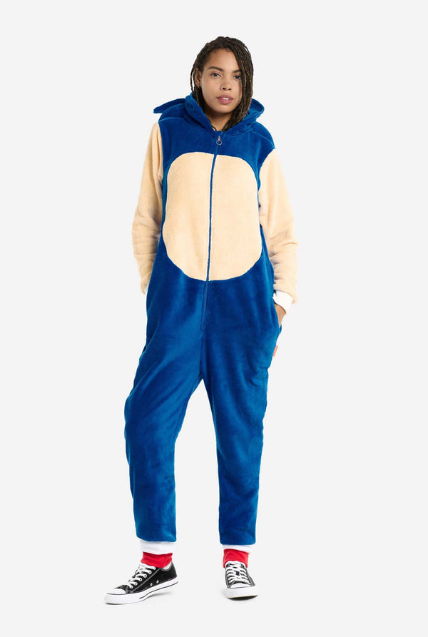 Een vrouw is gekleed in een gezellige, blauwe Sonic the Hedgehog onesie voor volwassenen, met zachtbeige patches op de voorkant en mouwen om de iconische look van Sonic na te bootsen. De onesie heeft een capuchon ontworpen met het herkenbare gezicht van Sonic. De ritssluiting aan de voorkant zorgt voor een comfortabele pasvorm en de vrouw staat met haar handen in de zijzakken van de onesie. Haar uitdrukking is kalm en ontspannen. 