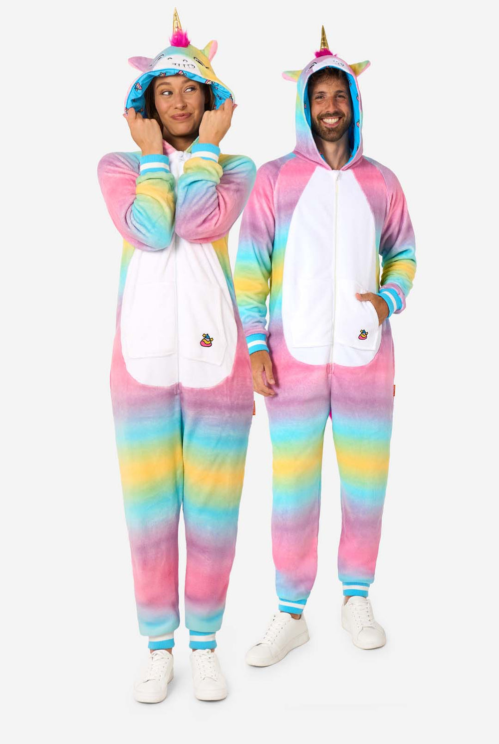 Een man en vrouw dragen een kleurrijke onesie voor volwassenen ontworpen als een regenboog eenhoorn. De onesie heeft een levendig verloop van pastelkleuren, een gouden hoorn en speelse eenhoorndetails, perfect voor gezellig loungen of verkleedfeestjes. De mannen en vrouwen hebben allebei hun capuchon op.