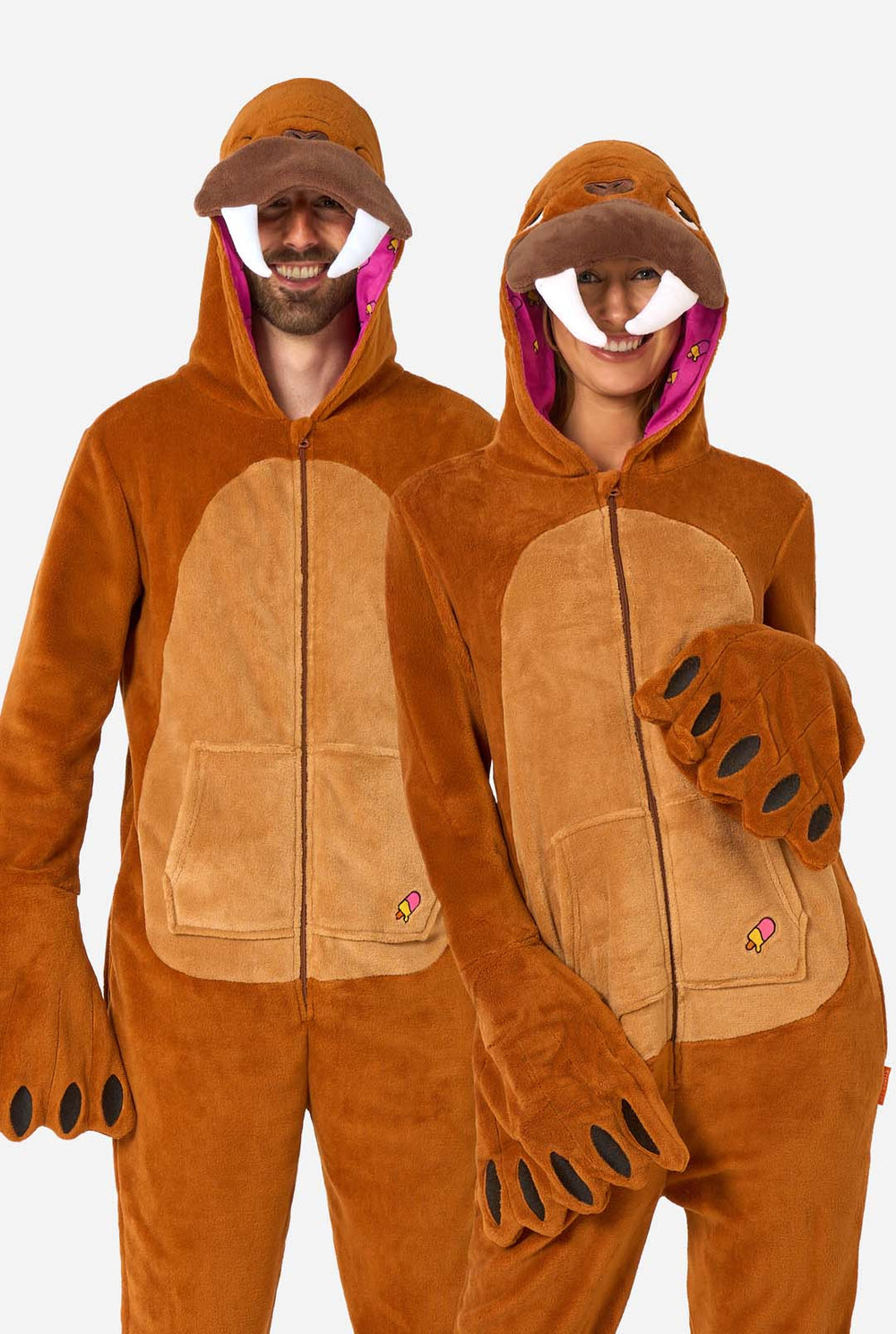 Een vrolijke man en vrouw dragen een bruine onesie voor volwassenen die is ontworpen als walrus. De onesie heeft een capuchon met hoektanden, opgestikte handschoenen die op poten lijken en speelse walrusdetails, perfect voor gezellig loungen of verkleed feestjes. De capuchon zit op hun hoofd.