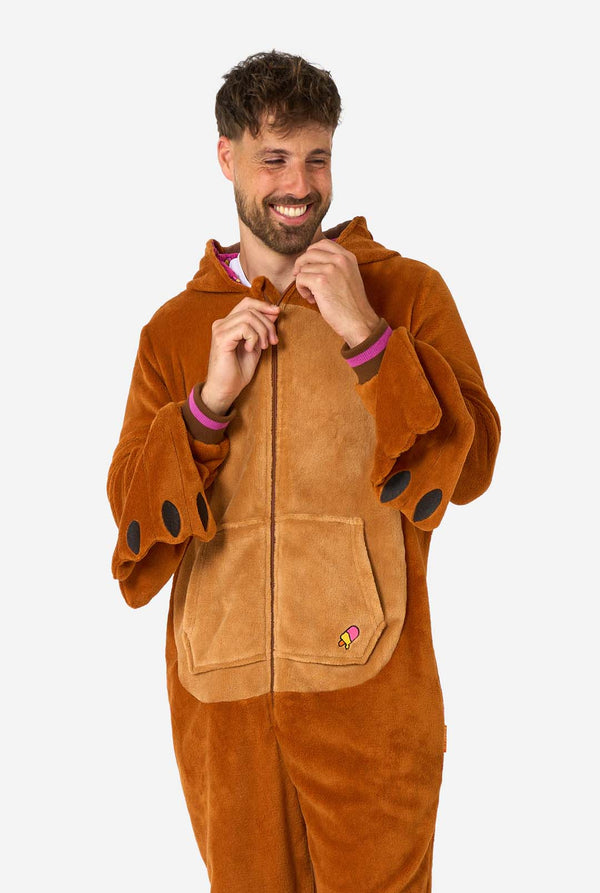 Een vrolijke man in een bruine volwassen onesie uitgevoerd in de vorm van een walrus. De onesie heeft een capuchon met hoektanden, gestikte handschoenen die op poten lijken en speelse walrusdetails, perfect voor gezellig loungen of verkleedfeestjes. De capuchon hangt op zijn rug.