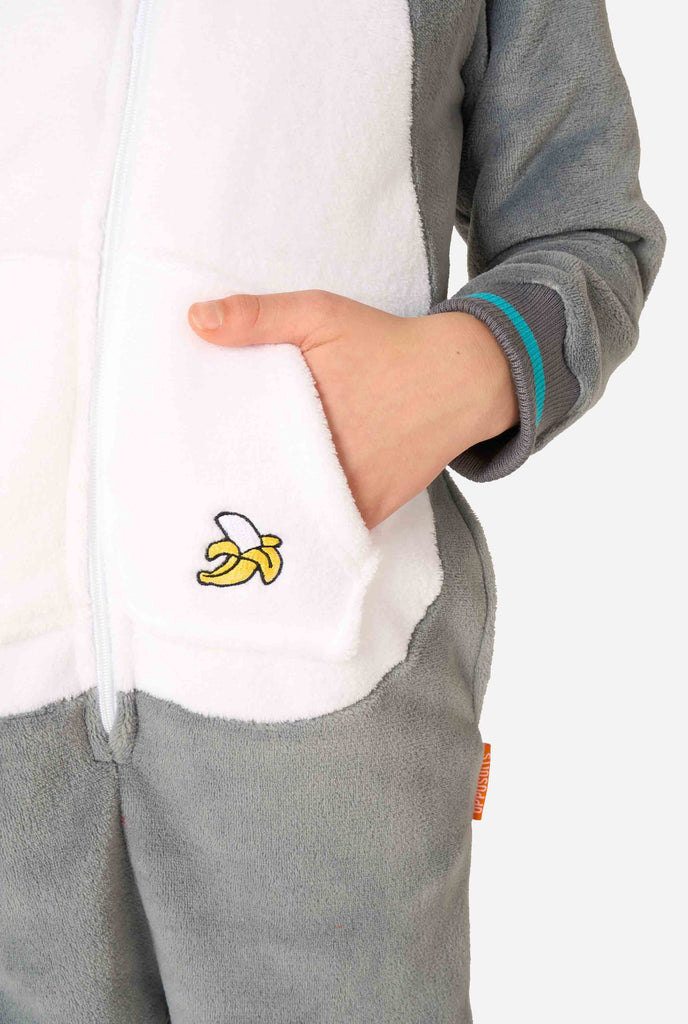 Een kind draagt ​​een comfortabel unisex kinderonesie in de vorm van een maki. De stof is gemaakt van grijs en wit pluche, heeft een capuchon met een cartoonmakigezicht en -oren en een speelse bananenprint op de zak. Gefocust op een hand in een zak.