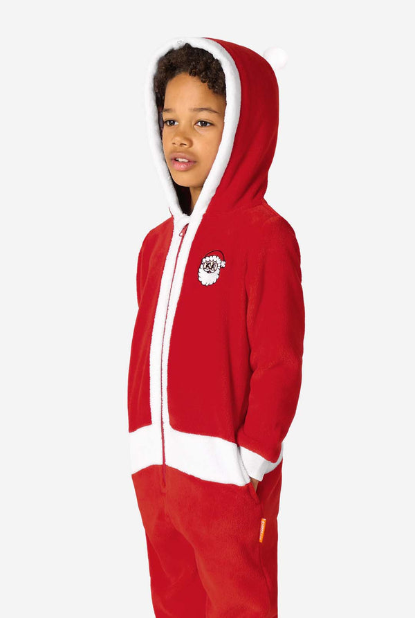 Kind dat een rode Santa-onesie met een wit afgewerkte capuchon draagt, iets zijwaarts staand met de handen in de zakken.