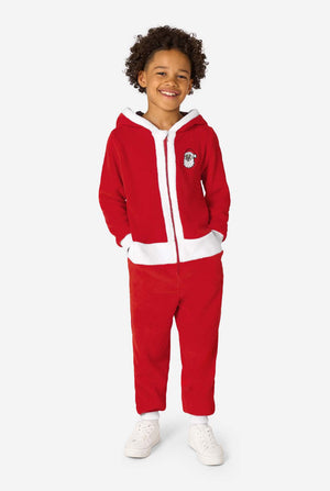 Volledige lichaamsweergave van een kind dat staat en glimlacht in een rode Santa-onesie met witte afwerking en witte sneakers.