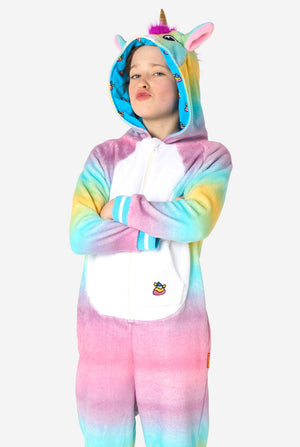 Een lachend kind draagt ​​een witte, roze, blauwe en gele kinderonesie ontworpen als eenhoorn. De Unicorn onesie heeft een capuchon die lijkt op een eenhoornkop en andere speelse details