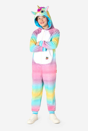 Een lachend kind draagt ​​een witte, roze, blauwe en gele kinderonesie ontworpen als eenhoorn. De Unicorn onesie heeft een capuchon die lijkt op een eenhoornkop en andere speelse details. Volledig lichaamsbeeld.