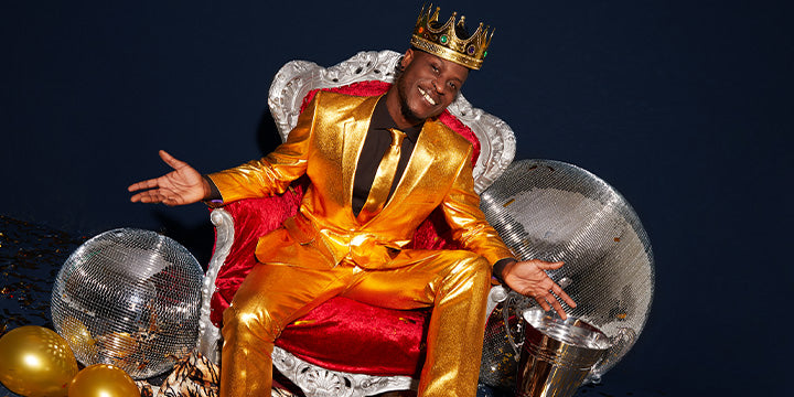 Man in a gold suit and crown sitting on a throne with disco balls and balloons.