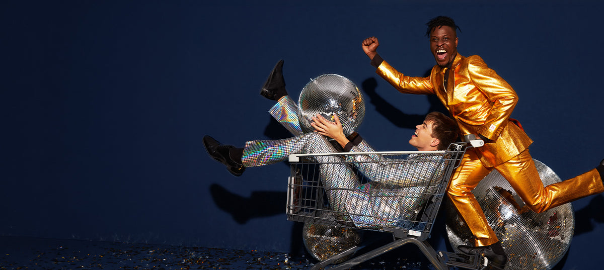 Two men in shiny party suits: one in gold pushes a shopping cart carrying a laughing man in a holographic mirror-ball suit.