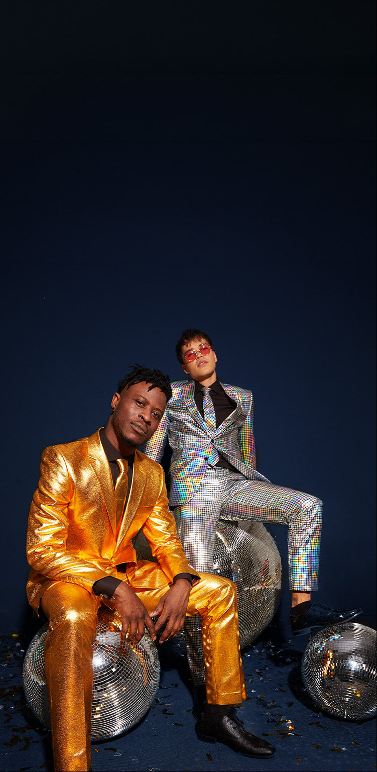 Two men in shiny party suits: one in gold pushes a shopping cart carrying a laughing man in a holographic mirror-ball suit.
